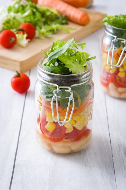 Foto ensalada saludable en mason jar con judías blancas, tomates, cebolla, maíz y lechuga en la mesa de madera