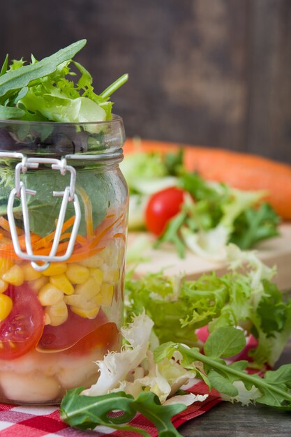 Foto ensalada saludable en mason jar con judías blancas, tomates, cebolla, maíz y lechuga en la mesa de madera