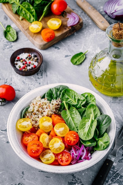 Ensalada de tazón verde saludable con espinacas, quinua, tomates amarillos y rojos, cebollas y semillas sobre fondo gris