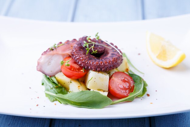 Ensalada tibia con pulpo, patatas, tomates cherry, rúcula, microgreens y limón en una placa blanca.