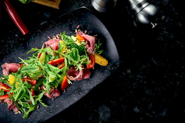 Ensalada tibia con rosbif y verduras frescas, servida en un plato negro sobre una mesa de mármol. Comida de restaurante. De cerca, enfoque selectivo