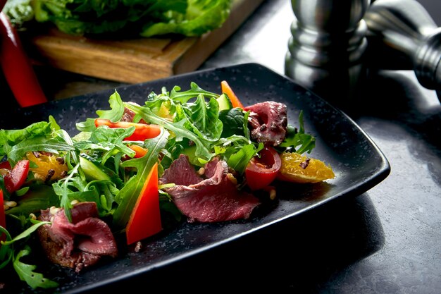 Ensalada tibia con rosbif y verduras frescas, servida en un plato negro sobre una mesa de mármol. Comida de restaurante. De cerca, enfoque selectivo