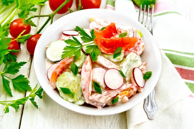 Ensalada de tomates frescos pepinos y rábanos con cebollas verdes y perejil aromatizados con mayonesa y crema agria en una toalla de plato y tenedor sobre el fondo de una tabla de madera clara