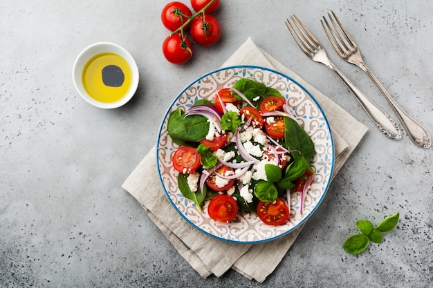 Ensalada de tomates, hojas de espinaca, cebollas rojas y queso feta en placa de cerámica ligera sobre fondo gris