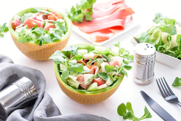 Ensalada tradicional italiana o española de melón fresco con prosciutto o jamón queso mozzarella y hierbas verdes en un plato de melón sobre una mesa blanca vista superior plato ligero de cocina mediterránea