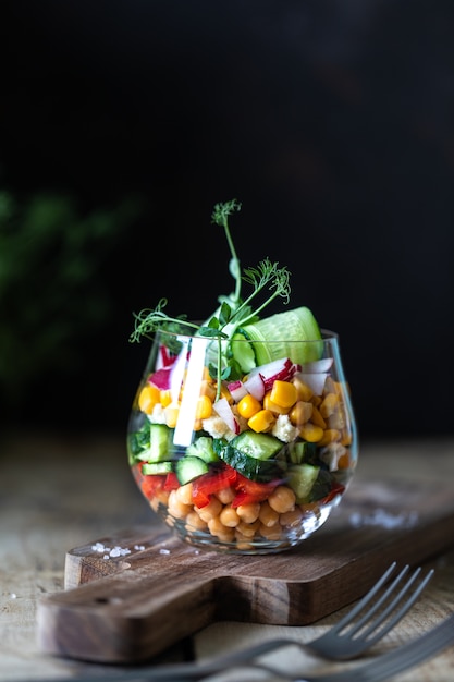 Ensalada vegetariana Arco iris en una taza de cristal sobre un fondo de madera. Nutrición apropiada. Copia espacio Vertical. Oscuro