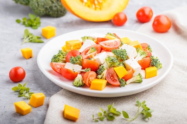 Ensalada vegetariana con brócoli, tomate, queso feta y calabaza en plato de cerámica blanca sobre una superficie de hormigón gris, vista lateral, de cerca.