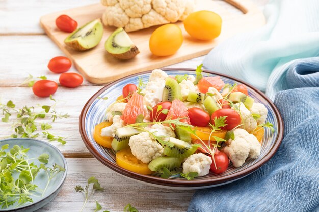 Ensalada vegetariana de col coliflor, kiwi, tomates, brotes microverdes sobre fondo blanco de madera