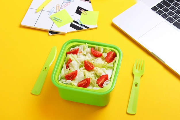 Ensalada vegetariana fresca en fiambrera verde con tenedor y cuchillo de plástico en el lugar de trabajo de la oficina. Cerca de merienda saludable en recipiente de plástico. Concepto de comida saludable. Vista superior, endecha plana, fondo amarillo.