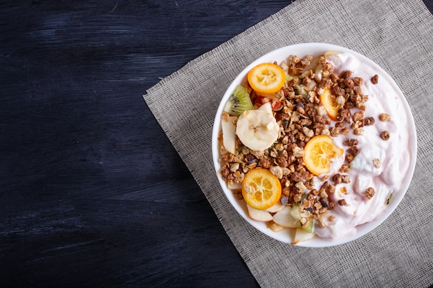 Ensalada vegetariana de plátanos, manzanas, peras, kumquats, kiwi con granola y yogur en madera negra