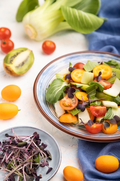 Ensalada vegetariana de repollo pac choi, kiwi, tomates, kumquat, brotes microverdes sobre un fondo de hormigón blanco y textil de lino azul. Vista lateral, primer plano, enfoque selectivo.