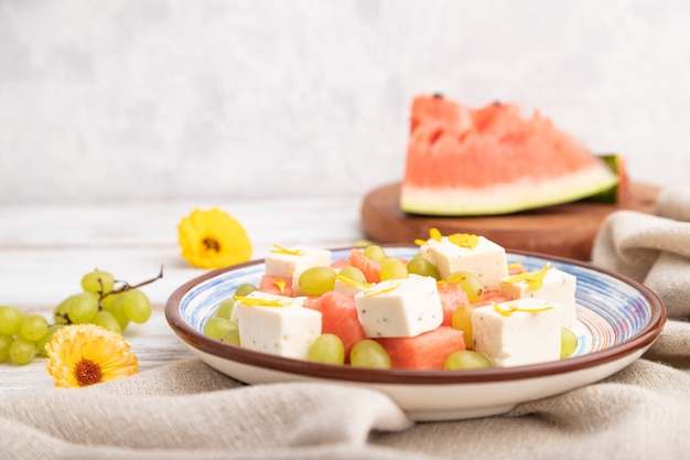 Ensalada vegetariana con sandía, queso feta y uvas en placa de cerámica azul sobre fondo blanco de madera y textiles de lino. Vista lateral, primer plano, enfoque selectivo.