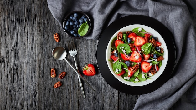 Ensalada de verano de fresas, arándanos, espinacas, nueces pecanas y queso feta desmenuzado en un recipiente blanco sobre una mesa de madera oscura con un paño gris, vista horizontal desde arriba, plano, espacio libre