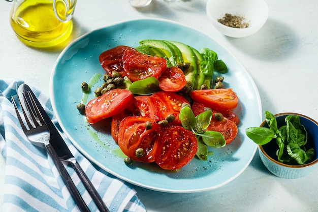 Ensalada de verano con tomates maduros y aguacate con alcaparras en un plato azul sobre la mesa