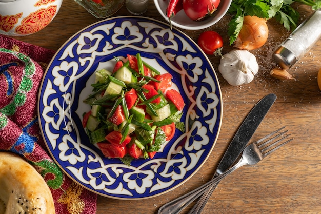 Foto ensalada de verano vegano vegetal de tomates, pepinos, tomates cherry, menta y cebollas verdes, sazonada con aceite de oliva y jugo de limón en un plato con un patrón uzbeko