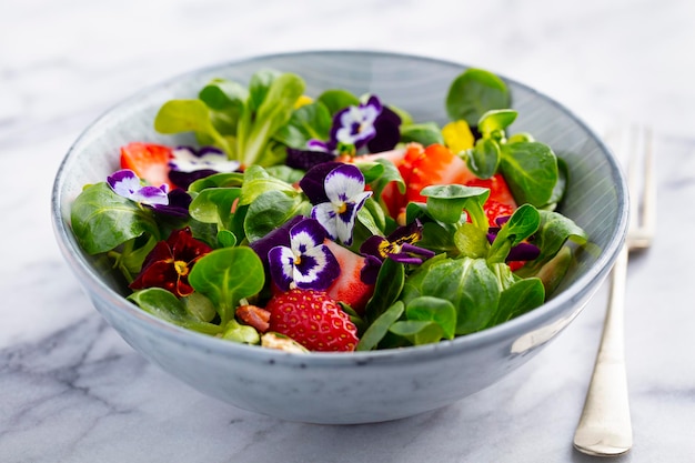 Ensalada verde fresca con fresas y flores comestibles en un bol Fondo de mármol Cerrar