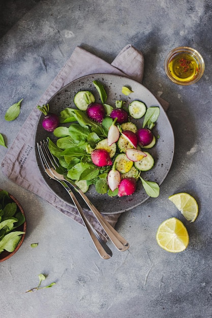 Ensalada verde con rúcula, pepino, rábano, lima, aceite de oliva, hierbas aromáticas sobre fondo de piedra gris