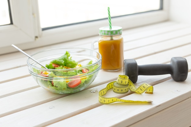 Ensalada de verduras y batidos de frutas y mancuernas se encuentran en un alféizar blanco. Concepto de actividad física de estilo de vida saludable y nutrición adecuada.