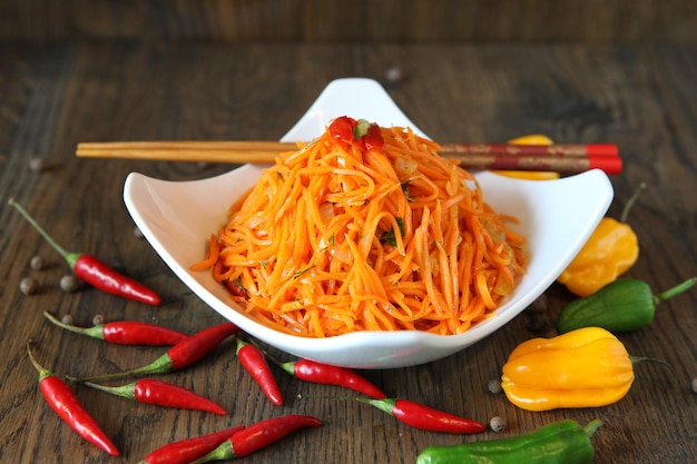 Ensalada de verduras coreana de zanahoria en un recipiente con palillos en la mesa de madera de roble y hojas de ensalada verde, pimientos rojos frescos. Ensalada picante de zanahoria al estilo coreano sobre un fondo oscuro. Cocina de Koryo-saram.