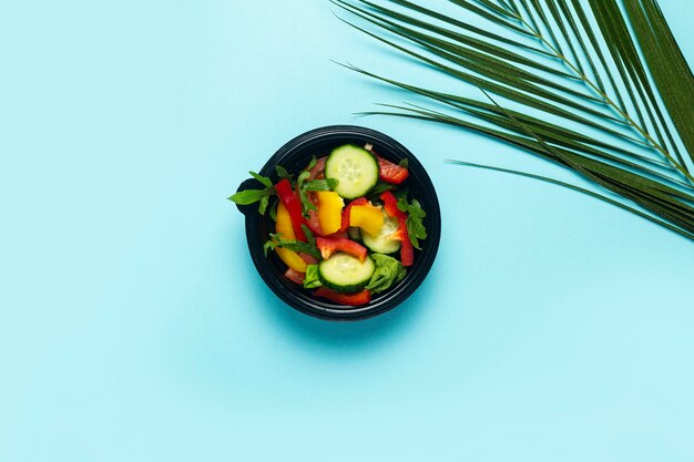 Ensalada de verduras en un recipiente de plástico desechable y una hoja verde de una palmera sobre un fondo azul. Vista superior, endecha plana.