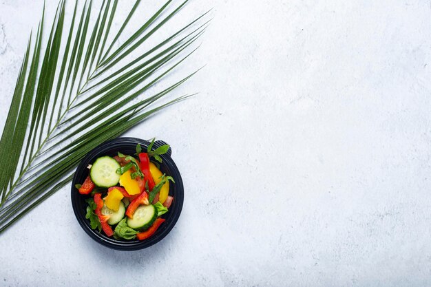 Ensalada de verduras en un recipiente de plástico desechable y una hoja verde de una palmera sobre un fondo biton.