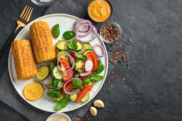 Ensalada de verduras con tomates, pepinos y rábanos y cabezas de maíz en un plato plano blanco sobre una pared negra con espacio para copiar. El concepto de comida vegetariana.