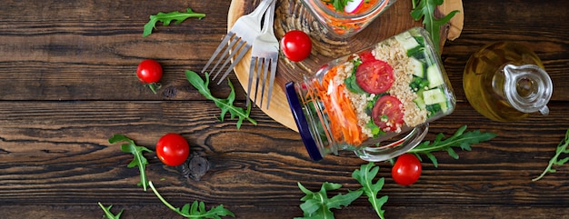 Ensaladas con quinua, rúcula, rábano, tomate y pepino en frascos de vidrio sobre mesa de madera. Comida sana, dieta, desintoxicación y concepto vegetariano. Vista superior. Lay Flat