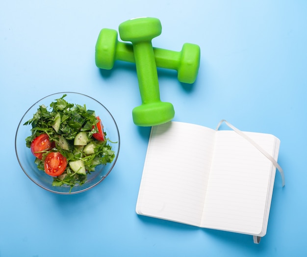 Ensaladera fresca, pesas y diario de fitness con páginas en blanco sobre fondo azul brillante