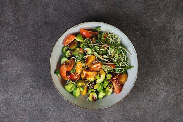 Ensaladera vegana con tomate, aguacate, pepino, microgreens y salsa de soja sobre un fondo oscuro