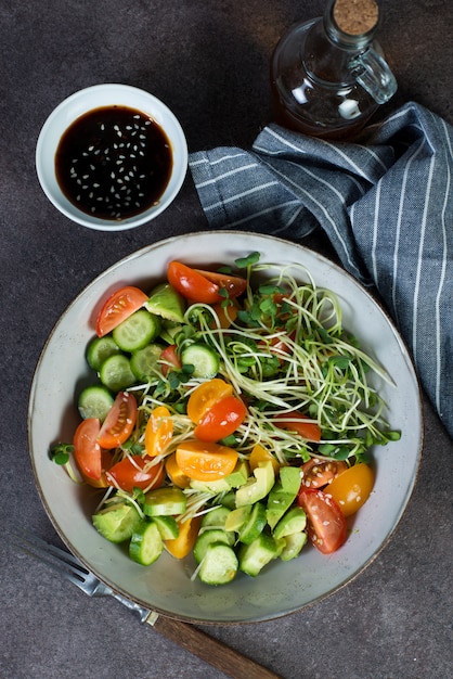 Ensaladera vegana con tomate, aguacate, pepino y microgreens sobre un fondo oscuro