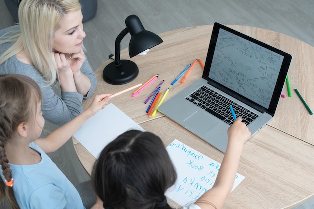 Enseñar en casa a dos hijas pequeñas que aprenden Internet en línea hacen la tarea usando la computadora con la ayuda de la madre, enseñan y alientan. Niña y hermana pequeña felices de estudiar educación en casa juntas.