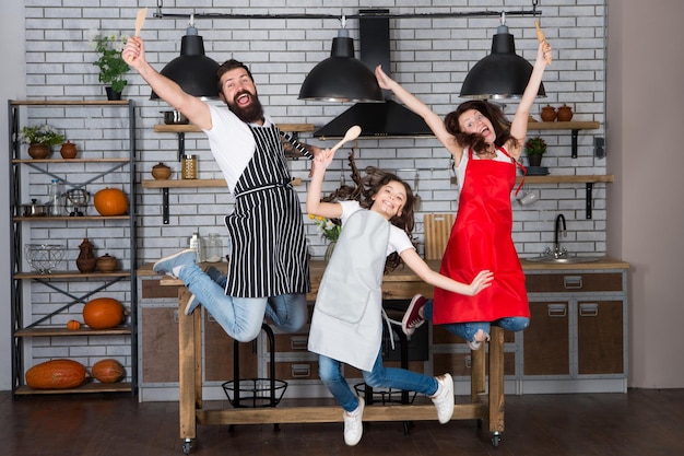 Ensine a criança a cozinhar. Café da manhã de fim de semana. Cozinhar com criança pode ser divertido. Se divertindo na cozinha. Família mãe pai e filha usam aventais saltam na cozinha. Família se divertindo cozinhando juntos.