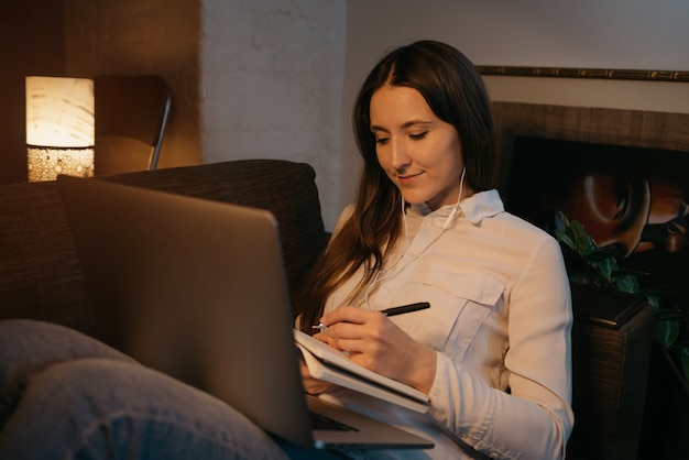 Ensino à distância. Uma mulher caucasiana com fones de ouvido, estudando remotamente em seu laptop. Uma garota bonita, fazendo anotações no caderno no sofá. Educação em casa.