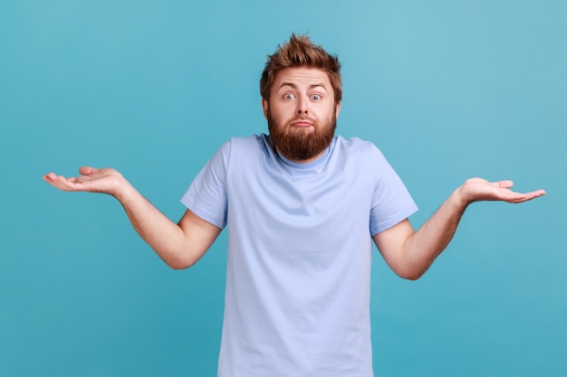 Então, quem se importa. Retrato de homem barbudo confuso espalha palmas com hesitação não tem ideia ou ideia fica intrigada, parecendo impotente para a câmera. Tiro de estúdio interior isolado sobre fundo azul.