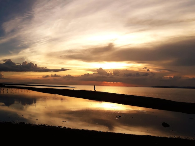 Entfernter Blick auf einen Silhouettenmann, der bei Sonnenuntergang am Strand gegen den Himmel spazieren geht
