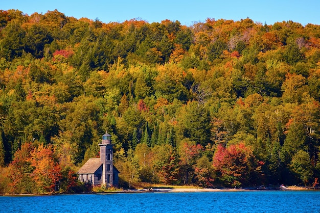 Entfernter Leuchtturm, umgeben von farbenfrohem Herbstwald und sandigem Ufer mit saphirblauem Seewasser