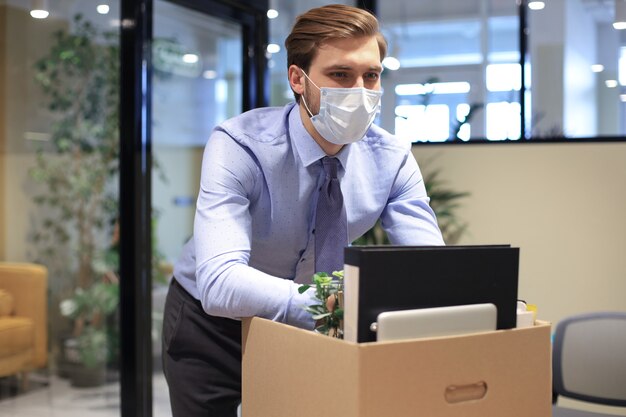 Entlassungsmitarbeiter in präventiver medizinischer Maske bei einem epidemischen Coronavirus. Trauriger entlassener Arbeiter nimmt seine Büromaterialien aus dem Büro mit.