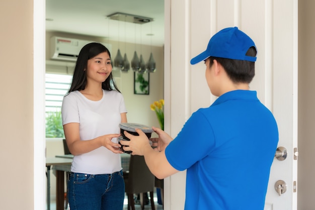 Entrega asiática joven en uniforme azul sonrisa y sosteniendo un montón de cajas de cartón en casa
