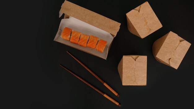 Entrega de comida asiática. Envases para sushi y woks. Alimentos en envases de papel sobre fondo negro. Paquete abierto con rollitos de salmón