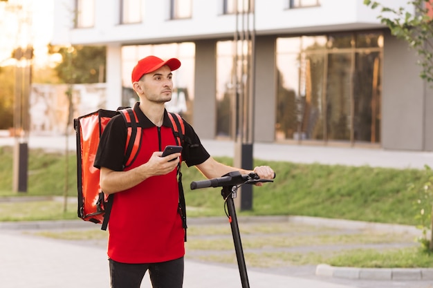 Entrega de comida de mensajero masculino con mochila térmica roja camina por la calle con scooter eléctrico utiliza phon