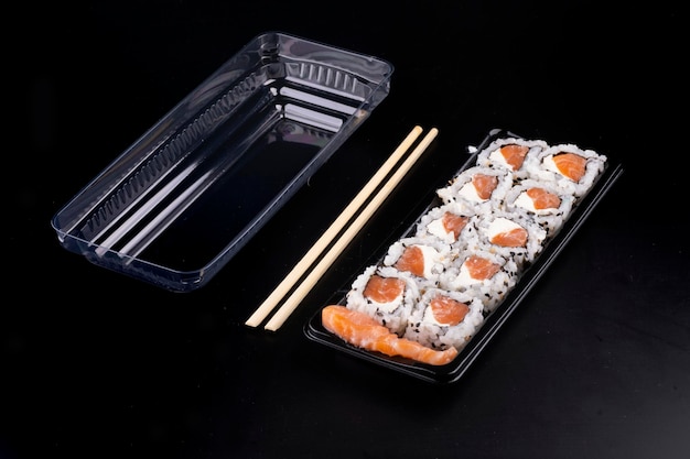 Entrega de comida oriental japonesa con envases de plástico abiertos y suhsis de salmón crudo con sashimi de queso crema y palillos sobre fondo negro en ángulo