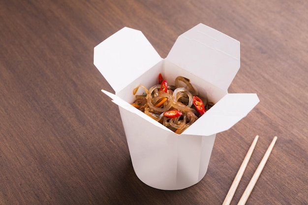 Foto entrega de comida de restaurante asiático. funchoza con carne y verduras, salsa de soja y guindilla en caja blanca para llevar y hashi sobre fondo de mesa de madera