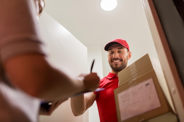 entrega, correio, pessoas e conceito de remessa - homem feliz com prancheta entregando caixas de encomendas para o cliente assinando formulário em casa