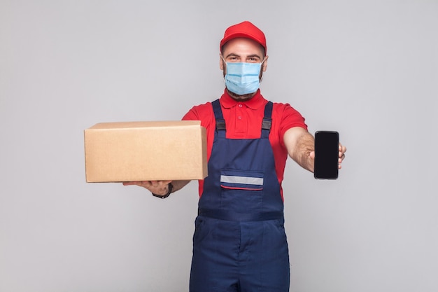 Entrega en cuarentena. ¡Esto es para ti! Hombre joven con máscara médica quirúrgica en uniforme azul y camiseta roja de pie, sosteniendo una caja de cartón y mostrando la pantalla del teléfono inteligente sobre fondo gris.