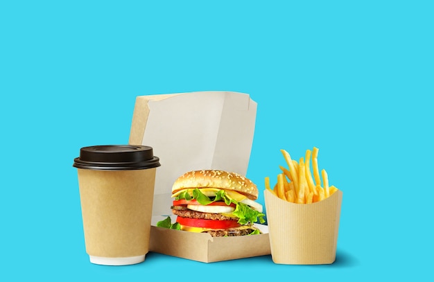 Entrega de almoço de fast food. Saboroso hambúrguer, batatas fritas e café em embalagem de papelão sobre fundo azul.