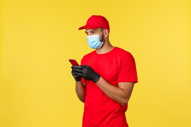 Entrega de comida, rastreamento de pedidos, conceito covid-19 e auto-quarentena. mensageiro ou funcionário em uniforme vermelho, máscara médica e luvas, usando smartphone, olhando para a esquerda, pesquisar o endereço do cliente para entregar