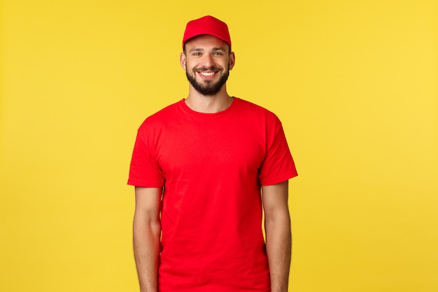 Entrega de correio expresso, transferência de pacotes e conceito de logística. Correio bonito e amigável com uniforme vermelho da empresa, camiseta e boné, sorrindo amplamente como um fundo amarelo de pé