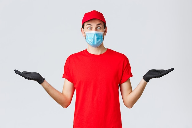 Entrega de mantimentos e pacotes, covid-19, quarentena e conceito de compras. correio surpreso em uniforme vermelho, luvas e máscara facial de mãos dadas lateralmente com banner ou promoção da empresa transportadora