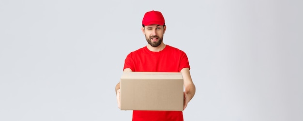 Entrega de pedidos on-line e conceito de envio de pacotes Correio barbudo bonito e atrevido em uniforme vermelho, entregando o pacote de caixa ao cliente O funcionário pisca para você e entrega o pacote do pedido