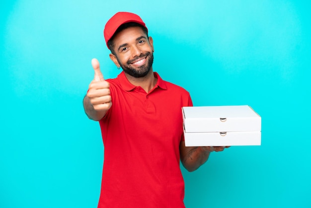 Entrega de pizza homem brasileiro com uniforme de trabalho pegando caixas de pizza isoladas em fundo azul com polegares para cima porque algo bom aconteceu